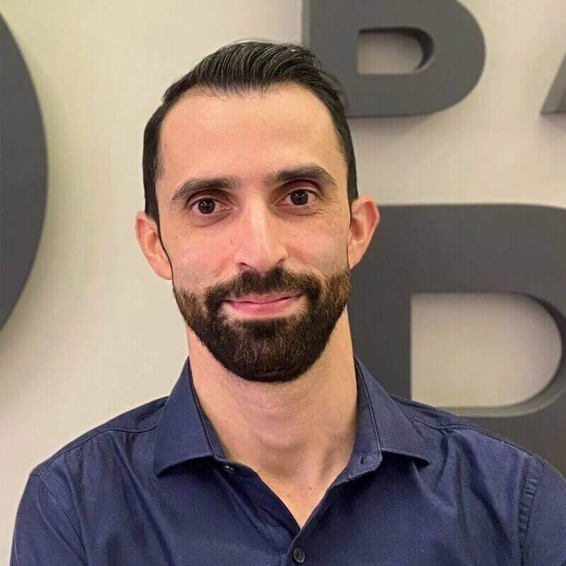 Homem branco de barba e cabelo escuro, usando camisa azul, posando em frente a uma parede com letras grandes.